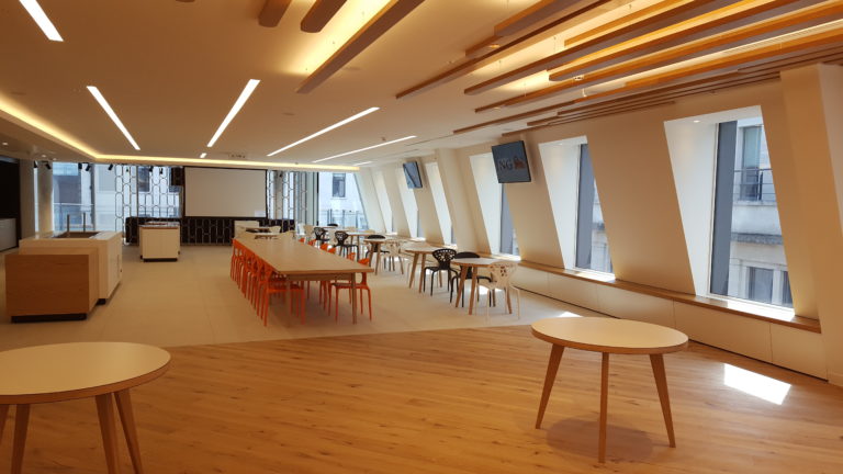 Spacious office break room with natural wood floors and a variety of seating options, including orange chairs and round tables. Overhead, linear lights complement the warm wooden ceiling slats. Large windows allow natural light to fill the room, and a monitor is mounted on the wall displaying a security camera feed.