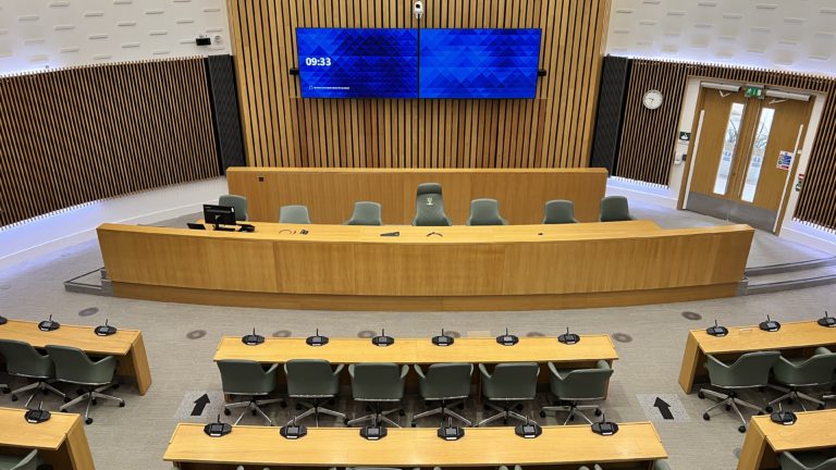 Interior of a modern council chamber with curved wooden desks and green chairs, facing a dais with more chairs and microphones. Two large digital screens showing '09:33' and a blue geometric pattern hang on the wood-panelled wall.