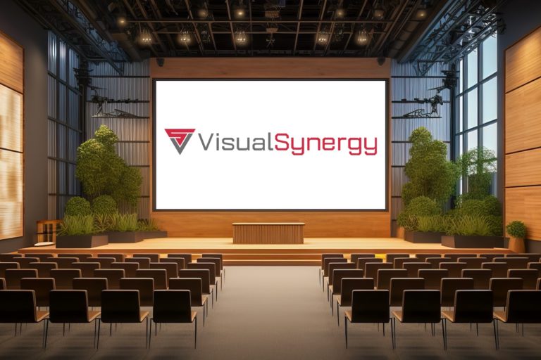 A modern conference room with rows of brown chairs facing a large screen displaying the logo of 'VisualSynergy' with a red and black colour theme. The room is well-lit, featuring wood paneling, green potted plants, and large windows with blinds partially down.